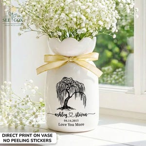 May include: A white ceramic vase with a scalloped rim, tied with a light gold ribbon. The front features a black graphic of a willow tree, the names "ashley" and "steven" with a heart, the date "04.14.2015", and the text "Love You More".