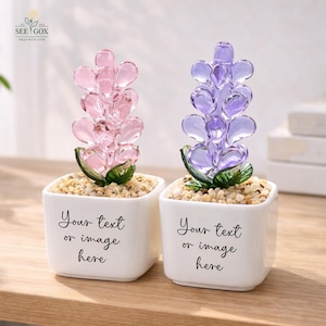 May include: Two square white ceramic pots filled with small pebbles, each containing a glass flower sculpture. One flower is light pink and the other is purple, both with green glass leaves. The pots feature the black text Your text or image here.