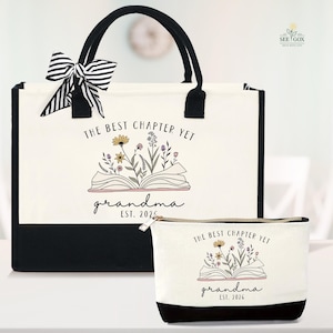 May include: A canvas tote bag and matching cosmetic bag. Both are off-white with black accents and feature the text "THE BEST CHAPTER YET grandma EST. 2026" with a floral illustration. The tote has black handles and a striped bow.