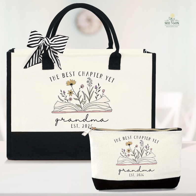 May include: A canvas tote bag and matching cosmetic bag. Both are off-white with black accents and feature the text "THE BEST CHAPTER YET grandma EST. 2026" with a floral illustration. The tote has black handles and a striped bow.