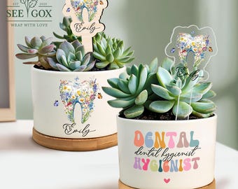 May include: Two white ceramic planters with succulents. One planter has a wooden tooth-shaped stake and the name "Emily". The other planter has the words "Dental Hygienist". Both planters sit on bamboo saucers.