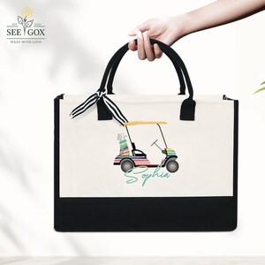 Puede incluir: Un bolso tote de lona con base y asas negras. El bolso presenta una colorida ilustraci&oacute;n de un carrito de golf con el nombre "Sophia" en fuente cursiva. La parte superior del bolso tiene una cinta a rayas blancas y negras atada con un lazo.