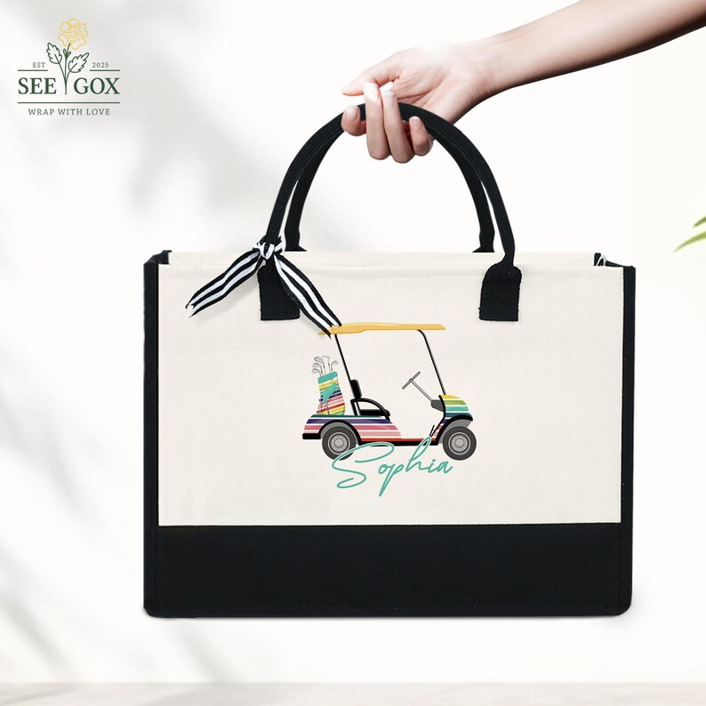 Puede incluir: Un bolso tote de lona con base y asas negras. El bolso presenta una colorida ilustraci&oacute;n de un carrito de golf con el nombre "Sophia" en fuente cursiva. La parte superior del bolso tiene una cinta a rayas blancas y negras atada con un lazo.
