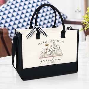 May include: A canvas tote bag with black handles and a black base. The bag features the text "THE BEST CHAPTER YET grandma EST. 2026" with an illustration of an open book and flowers. A black shoulder strap is attached.