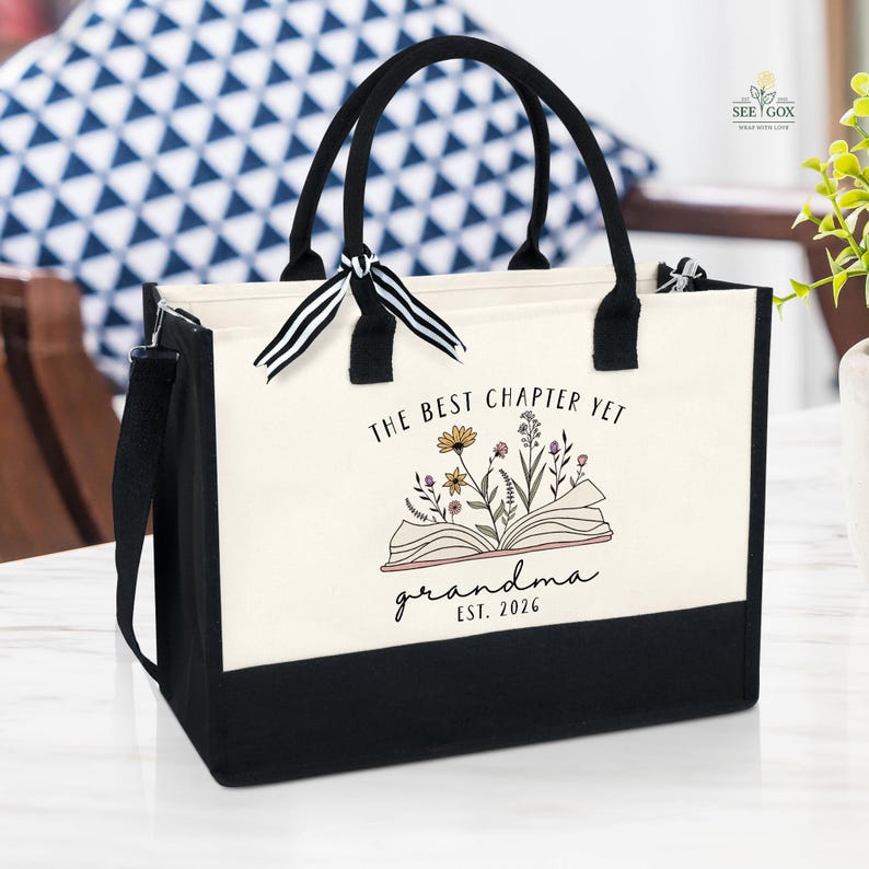 May include: A canvas tote bag with black handles and a black base. The bag features the text "THE BEST CHAPTER YET grandma EST. 2026" with an illustration of an open book and flowers. A black shoulder strap is attached.