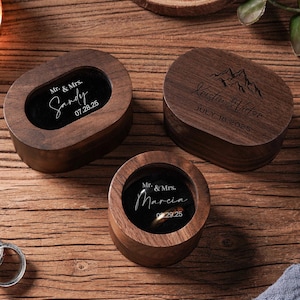 May include: Three wooden ring boxes with personalized inscriptions. One box is oval, two are round, and one is rectangular. The boxes are engraved with names and dates. Two silver rings are visible on the wooden surface.
