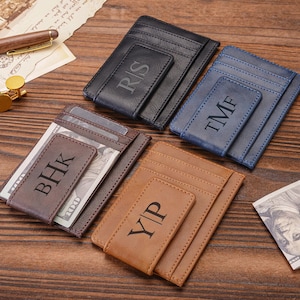 Puede incluir: Una variedad de billeteras con clip para dinero de cuero personalizadas en negro, azul, marrón y beige. Cada billetera presenta iniciales grabadas en la parte delantera. Una billetera está abierta, revelando un montón de billetes. Las billeteras están dispuestas sobre una superficie de madera.