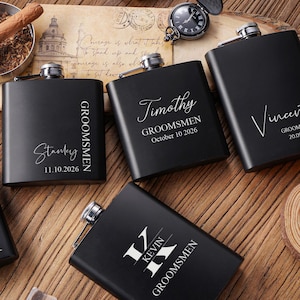 Könnte beinhalten: Schwarze Flachmänner mit silbernen Verschlüssen, personalisiert mit Namen und Daten. Die Flachmänner sind mit dem Wort "Groomsmen" und individuellen Namen graviert. Eine Zigarre und eine Taschenuhr befinden sich im Hintergrund.
