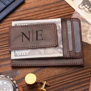 Può includere: Un portafoglio marrone in pelle con clip per banconote e le iniziali "NE" incise sul davanti. Il portafoglio contiene banconote da 100 euro e ha fessure per carte. Sono visibili anche un orologio, gemelli e una penna.