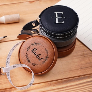 May include: A personalized leatherette tape measure with a white measuring tape extended. The tape measure is brown and black, with the name "Rachel" and the letter "E" engraved on the top. Pencils and a wooden tool are also visible.