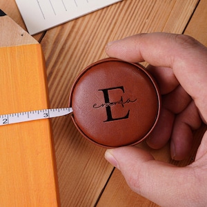 May include: A brown leatherette measuring tape with the letter "E" and the name "emanta" is held in a hand. A white measuring tape is extended across a large, orange pencil, measuring approximately 7.6 cm. The pencil has a black tip.