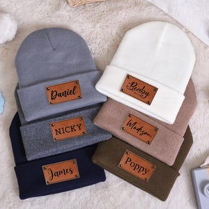 May include: A collection of personalized beanies in various colors, including gray, navy, white, and brown. Each beanie features a rectangular leather patch with a name engraved on it. The names include Daniel, Nicky, James, Becky, Madison, and Poppy.