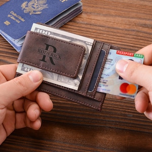 Puede incluir: Cartera de cuero marrón con un clip para billetes, que contiene un billete de 100 dólares. La cartera está personalizada con el nombre "Roderick" y una inicial. También se ve un pasaporte azul y una tarjeta de crédito.