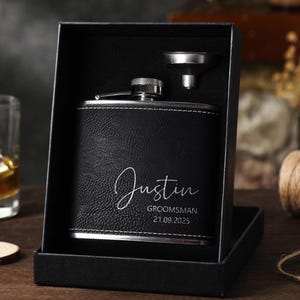 Puede incluir: Una petaca de acero inoxidable recubierta de cuero negro con costuras blancas y el texto grabado Justin, GROOMSMAN, 21.09.2025. La petaca se presenta en una caja de regalo negra con un pequeño embudo de metal plateado incluido para rellenar.