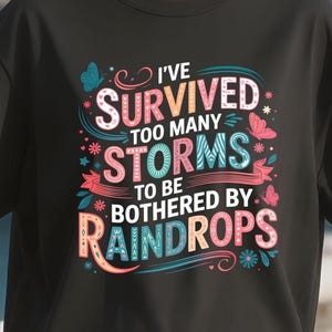 May include: Black t-shirt with a colorful graphic design. The design features the text "I've survived too many storms to be bothered by raindrops" in various fonts and colors, with butterfly and floral accents.