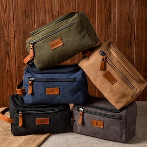 May include: A collection of five rectangular canvas toiletry bags in olive green, navy blue, tan, black, and charcoal grey. Each bag features a front zippered pocket, a main compartment with a zip, a side handle, and a brown leather patch with text.