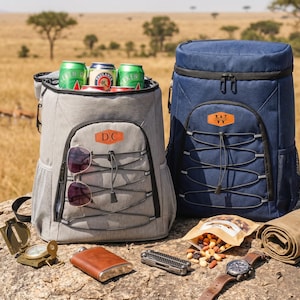 May include: A light gray and a navy blue backpack cooler, each featuring a front bungee cord system and a leather-like patch. The gray bag is open, revealing several beverage cans. Accessories include a compass, flask, multi-tool, watch, and snack bag.