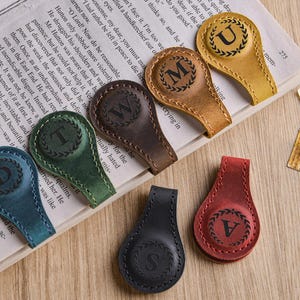 May include: A collection of leather bookmarks in various colors, including blue, green, brown, yellow, black, and red. Each bookmark features a different letter within a circular wreath design. They are resting on the pages of a book.