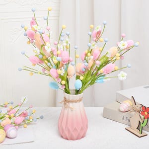 May include: A pink and white vase holds a bouquet of pastel-colored artificial flowers, including tulips and small round buds. Decorative Easter eggs and a wooden bunny figurine are also present, creating a festive spring arrangement.
