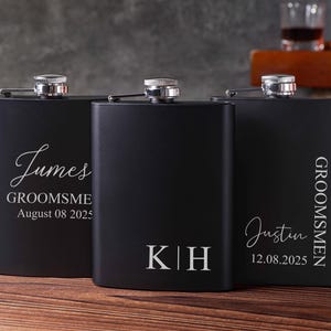 May include: Three black hip flasks with silver tops are arranged on a wooden surface. Each flask is engraved with a name, initials, and a date in a cursive font. The text includes the words "GROOMSMEN" and dates in the format DD.MM.YYYY.