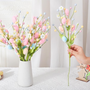 Könnte beinhalten: Eine weiße Vase mit pastellfarbenen Kunstblumen, darunter rosa Tulpen, blaue und gelbe Beeren und kleine weiße Blüten. Ein einzelner Stiel wird gehalten, und Ostereier liegen auf einem Teller.