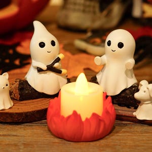 Peut inclure: Deux figurines de fantômes blancs assises sur des bûches sombres, l'une tenant un petit bâton marron, accompagnées de deux plus petites figurines blanches et d'une bougie centrale à pile avec une flamme jaune dans un support rouge.