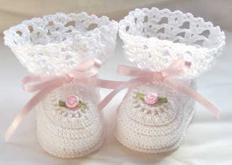 Puede incluir: Un par de patucos de beb&eacute; de ganchillo blanco con lazos de cinta de raso rosa y peque&ntilde;as rosas rosas. Los patucos tienen un delicado ribete de encaje alrededor de la parte superior.