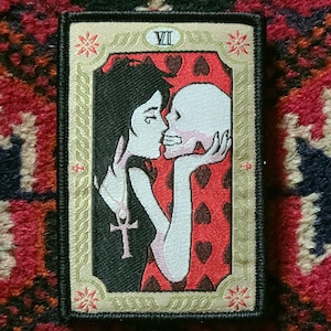 May include: An embroidered patch depicting a tarot card. The card is the Seven of Hearts and shows a woman kissing a skull. The card is framed in gold with a black border.