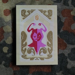 May include: Embroidered patch with a red and pink stylized figure, framed by ornate gold and cream details. The design is on a cream background, with a decorative border. The patch is rectangular.