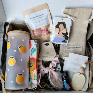 Peut inclure: Une boîte-cadeau remplie de divers articles, dont une bouteille d'eau en verre avec des points orange et blancs, un chouchou de couleur pêche, une cire fondue de couleur pêche avec le texte "Sun Kissed Peaches", une petite bouteille d'huile essentielle, un diffuseur en bois et une paire de boucles d'oreilles de couleur pêche.