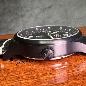 May include: A black wristwatch with a black dial and white numerals. The watch has a black bezel and a black crown with an airplane logo. The watch band is black leather with white stitching. The watch is on a wooden surface.