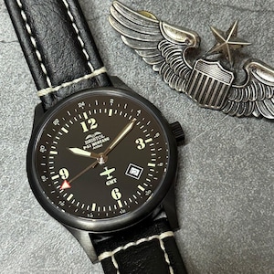May include: Black wristwatch with a black leather band and white stitching. The watch face is black with white numerals and hands. The watch has the text "BRISTOL P-51 MUSTANG Artifact" and a small airplane graphic. A silver metal pilot's wings pin is in the background.
