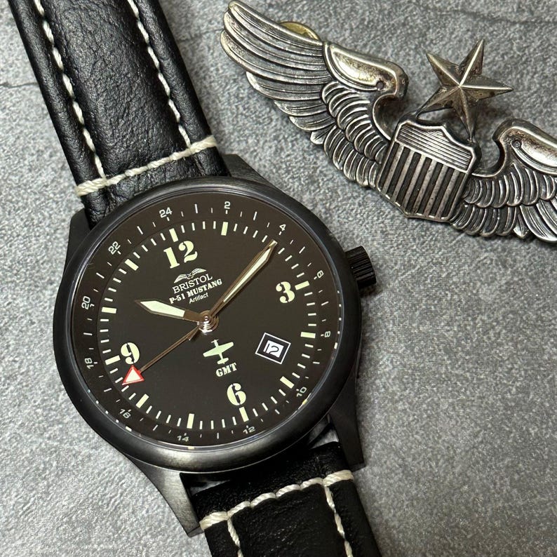 May include: Black wristwatch with a black leather band and white stitching. The watch face is black with white numerals and hands. The watch has the text "BRISTOL P-51 MUSTANG Artifact" and a small airplane graphic. A silver metal pilot's wings pin is in the background.