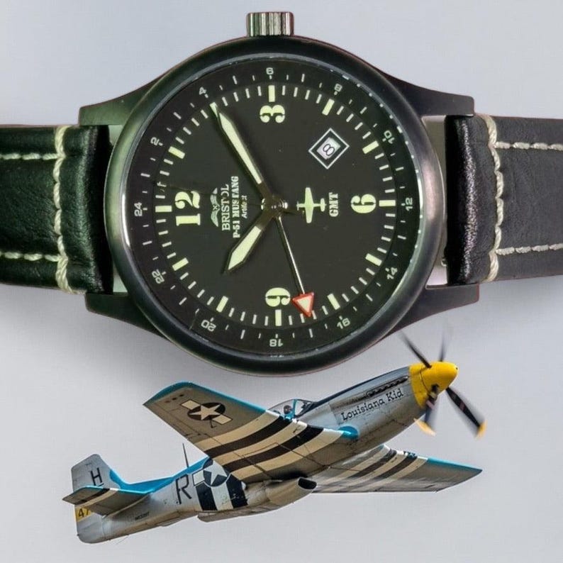 May include: A black wristwatch with a black leather band and white stitching. The watch face is black with white numerals and hands. Below the watch is a model airplane with the text "Louisiana Kid".