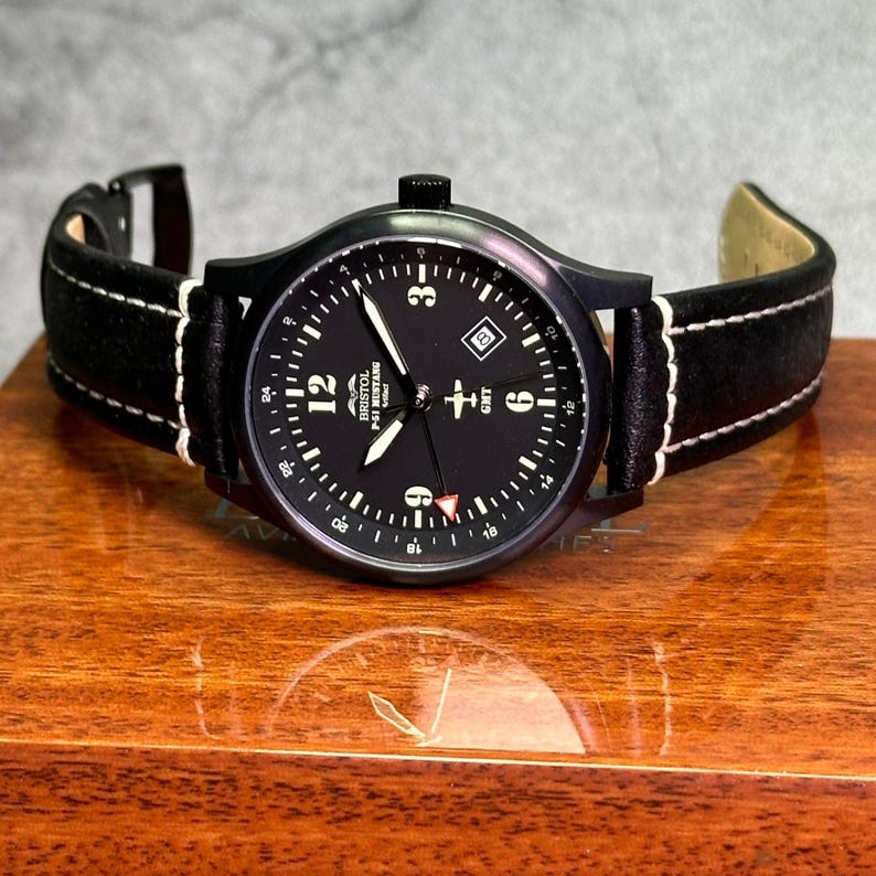 May include: A black wristwatch with a black face and a black leather band with white stitching. The watch face has white numerals and hands, and the text "BRISTOL P-51 MUSTANG" is visible. The watch is on a wooden surface.