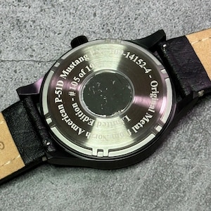 May include: Close-up of a black watch with a black leather strap. The watch back is visible, displaying text that reads "North American P-51D Mustang American Edition - #105 of 10 - Original Metal". The watch has a circular face.