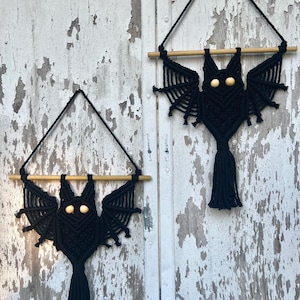 May include: Two black macrame bat wall hangings. Each bat has wooden bead eyes, a tassel tail, and is suspended from a wooden dowel and black string. The bats are displayed against a weathered, white-painted wooden background.