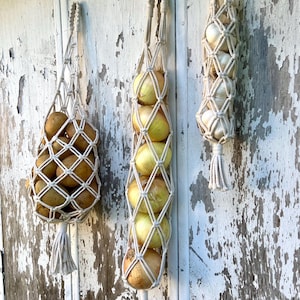 May include: Three macrame produce bags hanging on a distressed white wooden surface. One bag contains potatoes, another onions, and the third garlic. Each bag is made of off-white woven cotton with a looped handle and a tassel at the base.