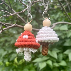 May include: Two macrame mushroom keychains. One mushroom is red with white spots, the other is white. Both have wooden beads and silver-colored clasps. The keychains are hanging from a tree branch against a blurred green background.