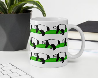 May include: White ceramic mug featuring a repeating pattern of cartoon pandas in black and white, lying on green stripes. The mug has a curved handle and is set against a neutral background with a plant and keyboard.