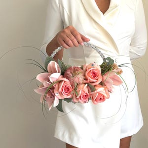 May include: A floral arrangement in a silver-toned, circular basket. The arrangement features pink roses, orchids, and other flowers, accented with greenery and crystals. The handle is embellished with rhinestones. The overall aesthetic is elegant and decorative.