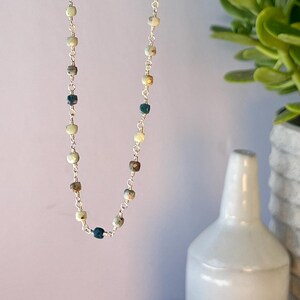 May include: A delicate necklace featuring small, multi-colored beads strung on a silver chain. The beads are a mix of blue, cream, and brown tones. The necklace is held up against a soft pink background, showcasing its length and design.