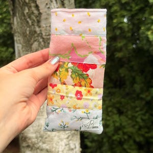 May include: A rectangular fabric pouch featuring a patchwork design of various floral and botanical patterns in shades of pink, yellow, green, and white. A small white fabric label with the black text EYBecca is sewn onto the bottom right corner.