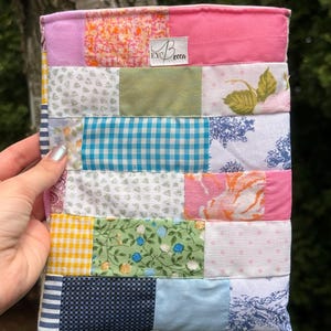 May include: A rectangular fabric pouch constructed from a patchwork of various cotton textiles featuring floral, gingham, polka dot, and solid patterns in shades of pink, blue, green, yellow, and white, with a white label reading Becca.
