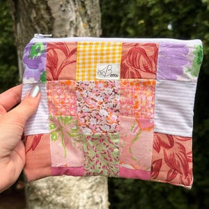 May include: A rectangular zippered pouch constructed from a patchwork of various fabrics in shades of pink, orange, yellow, and white. The design features floral, gingham, and striped patterns. A white label with the text "EYBecca" is sewn on the front.