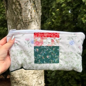 Peut inclure: Une pochette zippée en tissu rectangulaire présentant un motif patchwork avec des panneaux floraux blancs sur les côtés et une bande verticale centrale de tissu floral rouge, rose et vert foncé. Une étiquette blanche indique Becca.