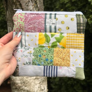 Puede incluir: Un estuche rectangular con cremallera hecho de un patchwork de varios cuadrados de tela con patrones florales, de cachemira, limones, cuadros y caras sonrientes en tonos amarillo, verde, blanco y rosa, con una etiqueta que dice EYBecca.