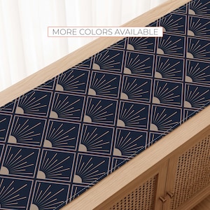 May include: A rectangular table runner featuring a dark navy blue background with a repeating geometric pattern of beige squares containing radiating sunburst lines. The text MORE COLORS AVAILABLE is printed in a white box at the top center.