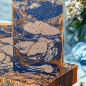 May include: Artisan soap bars with a marbled design. The soap features swirls of navy blue, tan, and orange. The bars are stacked, showcasing the intricate patterns and colors. The soap has a natural, handmade aesthetic.