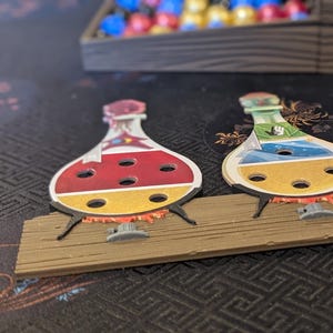 May include: A wooden game board with three colorful potion bottles. Each bottle has holes for balls to be dropped in. There are three balls on the board, one yellow, one red, and one blue.